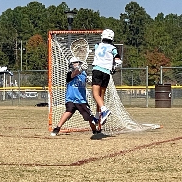 Youth lacrosse player jumps in front of a goal with a goalie defending.