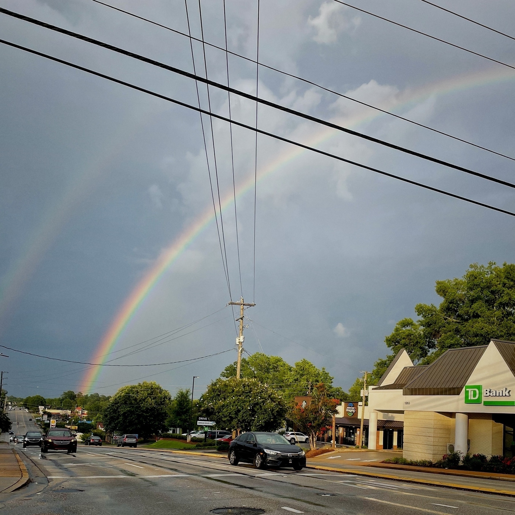 A vibrant double rainbow arches over a busy urban street lined with buildings and cars.